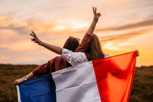 Two, young, females, covering themselves with flag of France. Standing on the meadow at sunset. Rear view.